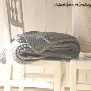 Handgestrickte Tagesdecke in Grau mit Perlmuster, gefaltet auf weißem Stuhl vor weißer Holzwand