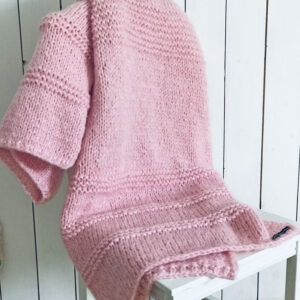 Handgestrickte Mohair Tagesdecke in Rosé, drapiert über weißem Stuhl vor Holzwand
