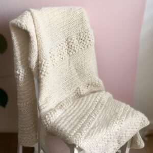 Handgestrickter Alpaka-Merino Überwurf in Weiß, drapiert auf weißem Stuhl vor rosé Wand