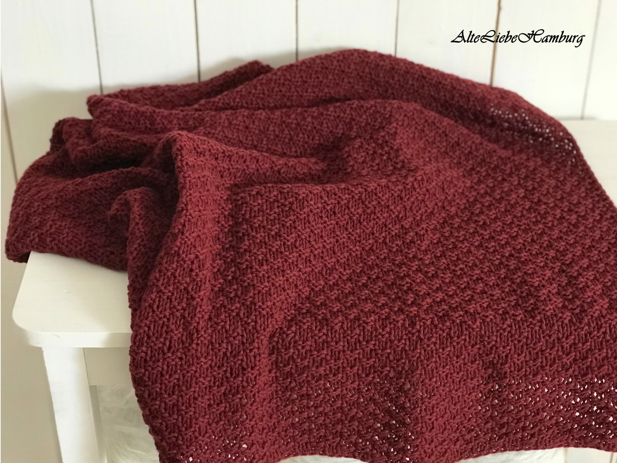 Handgestrickte Tagesdecke in Bordeaux-Rot mit Lochmuster-Rand, locker über eine weiße Bank drapiert – AlteLiebeHamburg