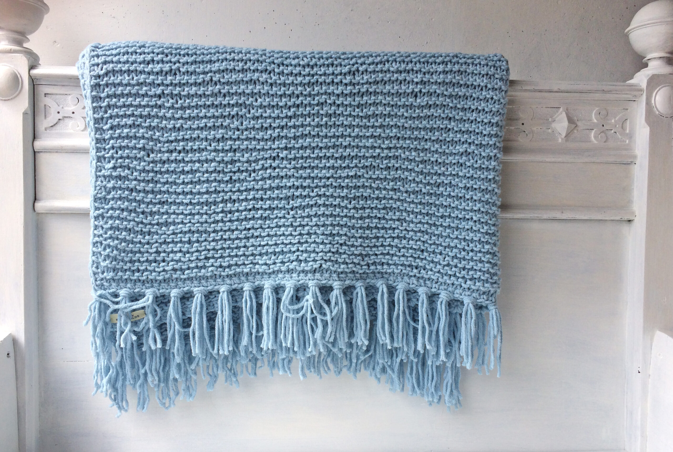 Handgestrickte Strickdecken mit Fransen in Blau, aufgereiht auf einem weißen Bettgestell – AlteLiebeHamburg
