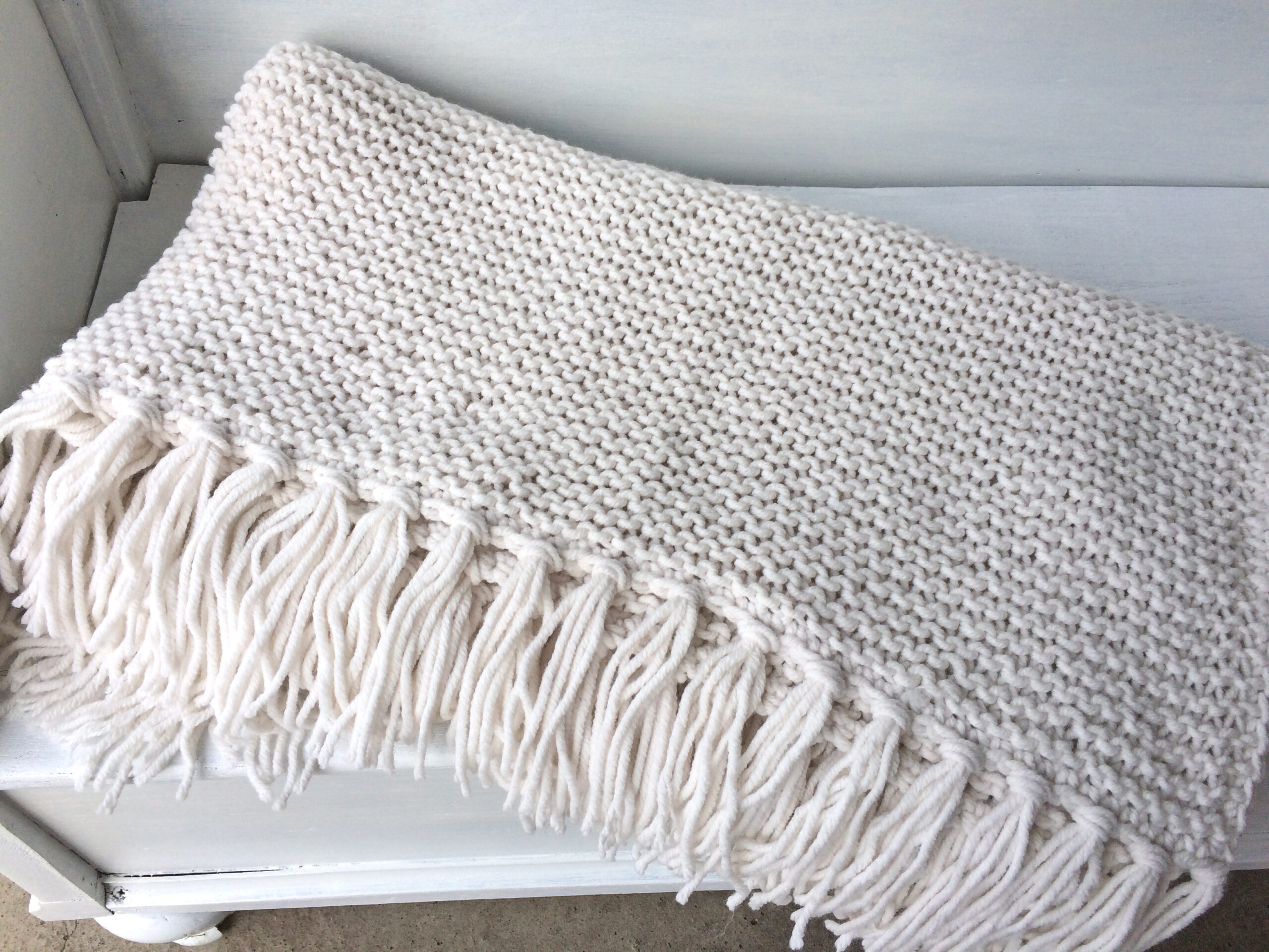 Handgestrickte Strickdecken mit Fransen in Weiß, aufgereiht auf einem weißen Bettgestell – AlteLiebeHamburg