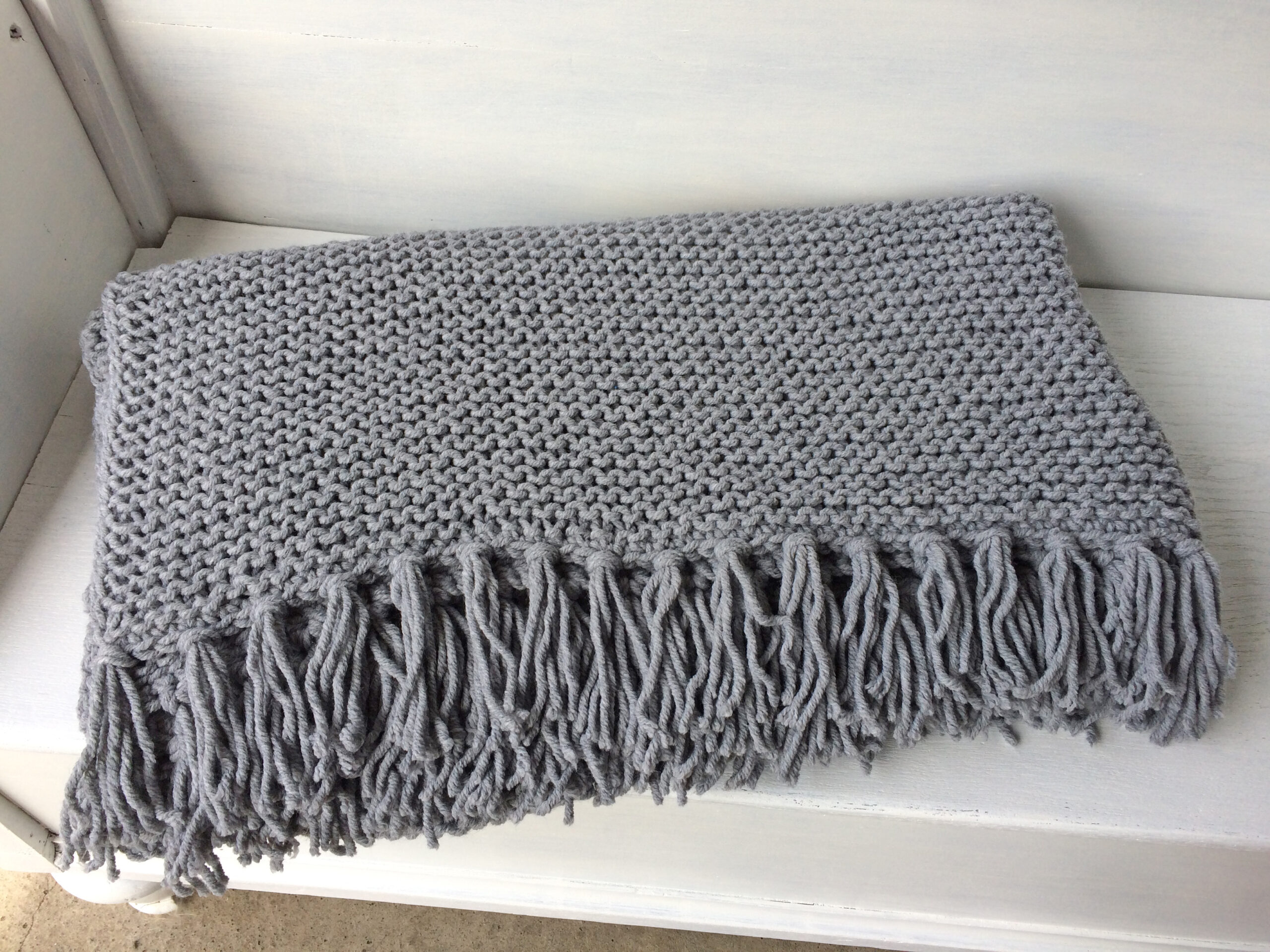 Handgestrickte Strickdecken mit Fransen in Grau, aufgereiht auf einem weißen Bettgestell – AlteLiebeHamburg