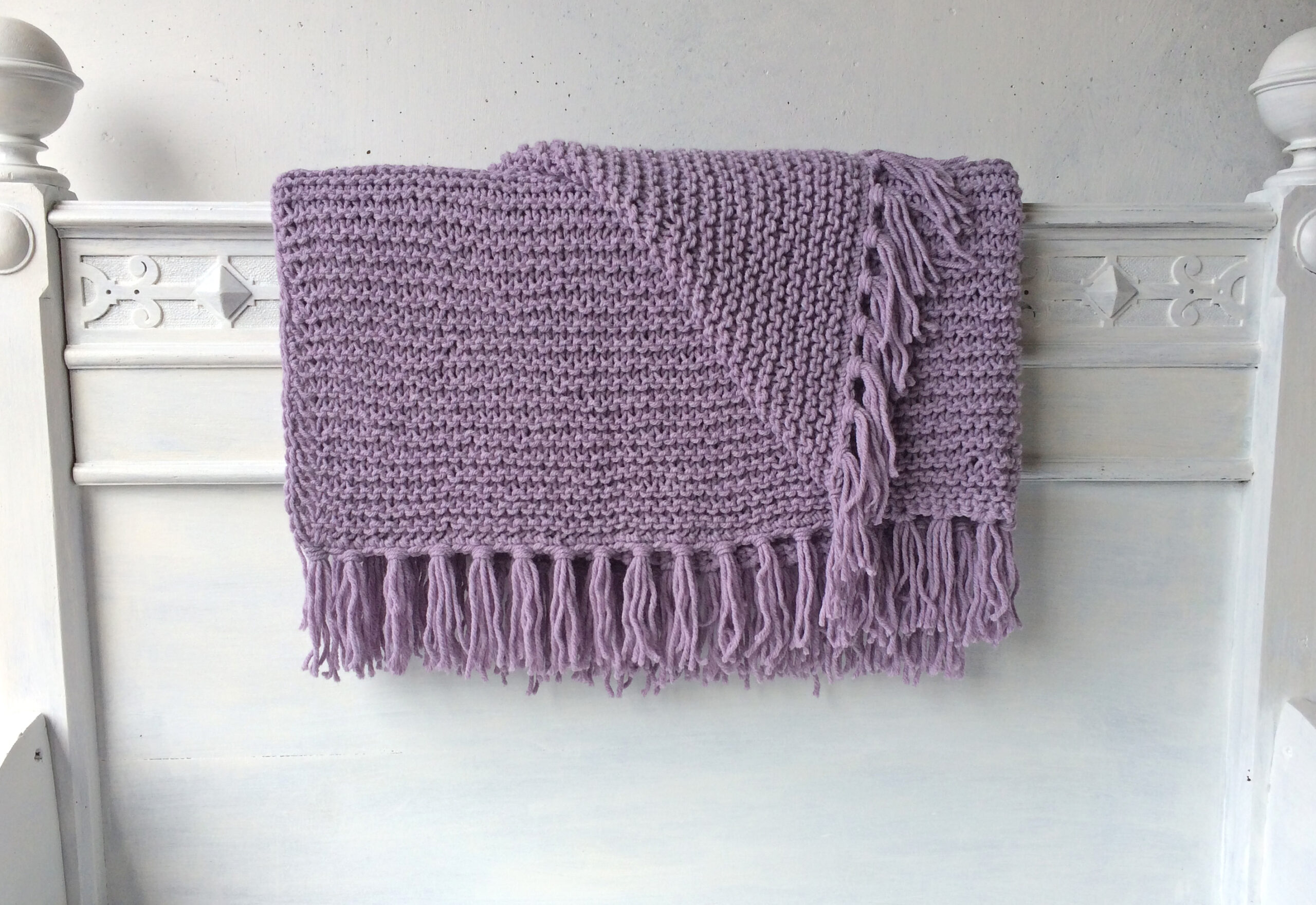 Handgestrickte Strickdecken mit Fransen in Flieder auf einem weißen Bettgestell – AlteLiebeHamburg
