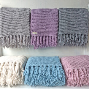 Handgestrickte Strickdecken mit Fransen in Grau, Flieder, Blau, Weiß und Rosé, aufgereiht auf einem weißen Bettgestell – AlteLiebeHamburg