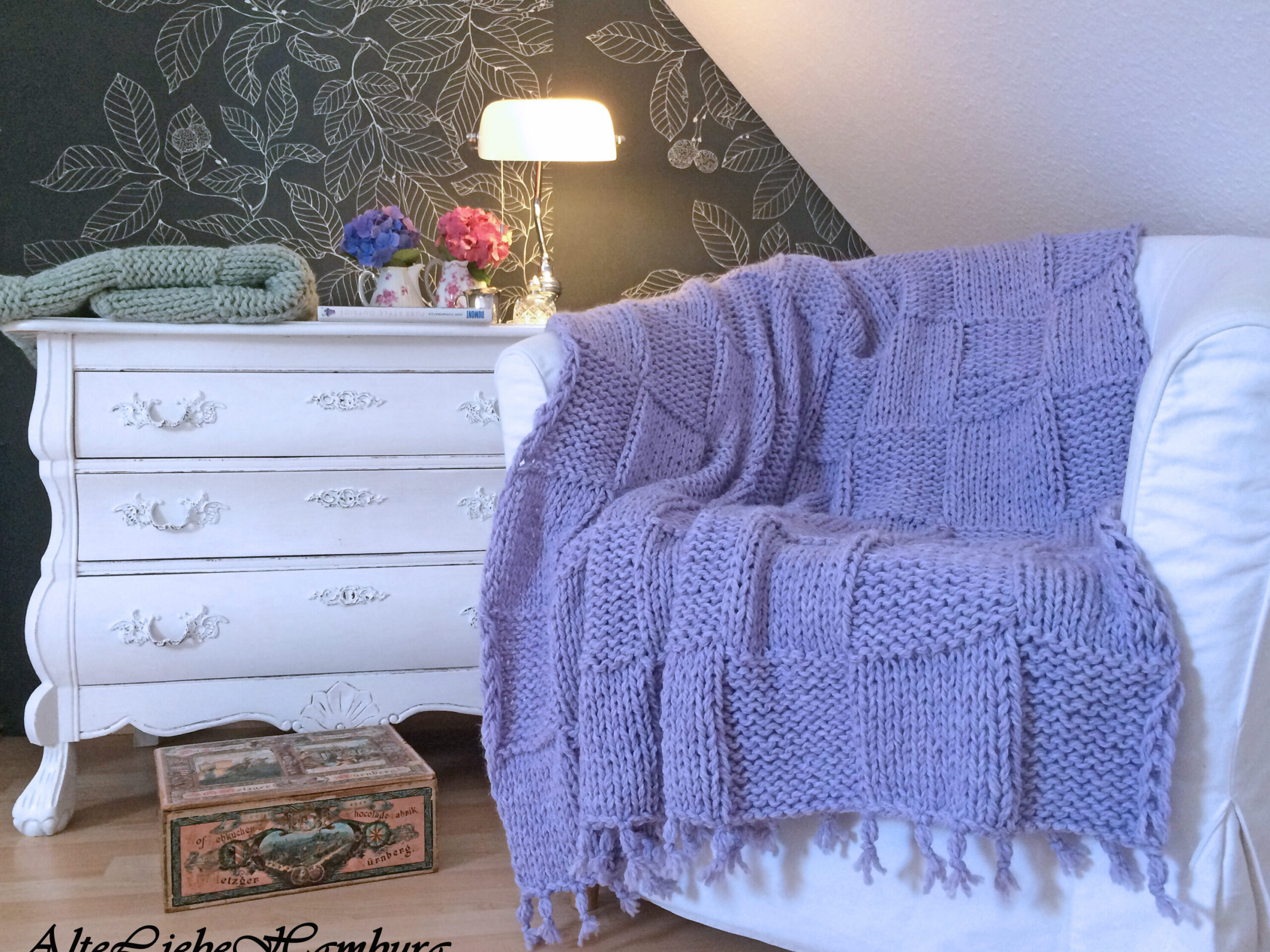 Cover der Strickanleitung für die Tagesdecke Shetland in Flieder, mit Foto der Decke auf weißer Kommode – AlteLiebeHamburg