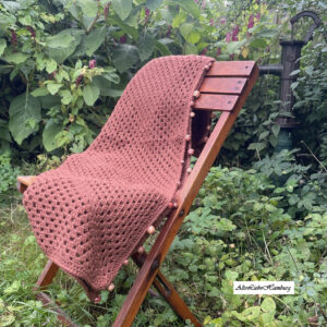 Handgehäkeltes Terracotta-Plaid mit Holzperlen, über eine Stuhllehne drapiert im Garten